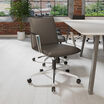 Modern office space with a sleek black ergonomic chair on casters, light wood flooring, large industrial windows, and minimalist furniture against an exposed brick wall backdrop.