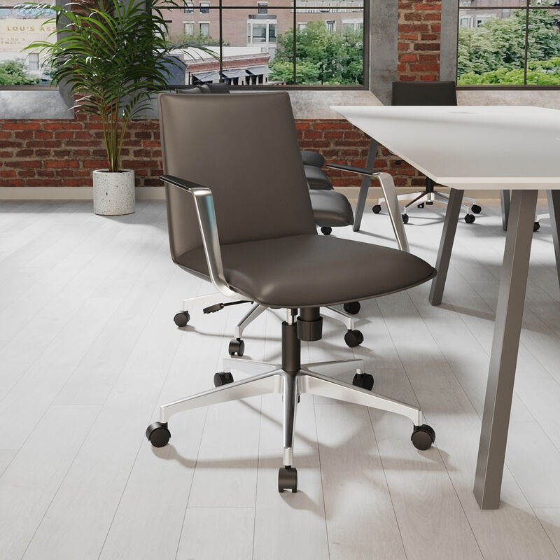Modern office space with a sleek black ergonomic chair on casters, light wood flooring, large industrial windows, and minimalist furniture against an exposed brick wall backdrop. image number null