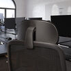 Close-up rear view of the black headrest mounted on a mesh office chair, showing the adjustable arm and mounting bracket, in an open office setting.