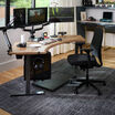 Modern home office setup featuring a black ergonomic Vari Tempur-Pedic chair at a curved wood desk with triple monitor arms, editing equipment, camera gear, and a gray area rug on hardwood flooring.