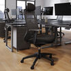 Front angled view of a black mesh task chair with an adjustable headrest, armrests, and a five-star caster base, shown in an open office with desks and monitors in the background.