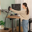 A woman stands at a height-adjustable standing desk in a home office, adjusting the Vari Studio Lamp angle.
