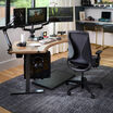 Modern home office setup featuring a dark gray ergonomic Tempur-Pedic chair at a curved wood desk with triple monitor arms, editing equipment, camera gear, and a gray area rug on hardwood flooring.