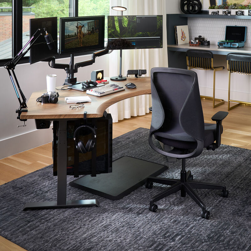 Modern home office setup featuring a dark gray ergonomic Tempur-Pedic chair at a curved wood desk with triple monitor arms, editing equipment, camera gear, and a gray area rug on hardwood flooring. image number null