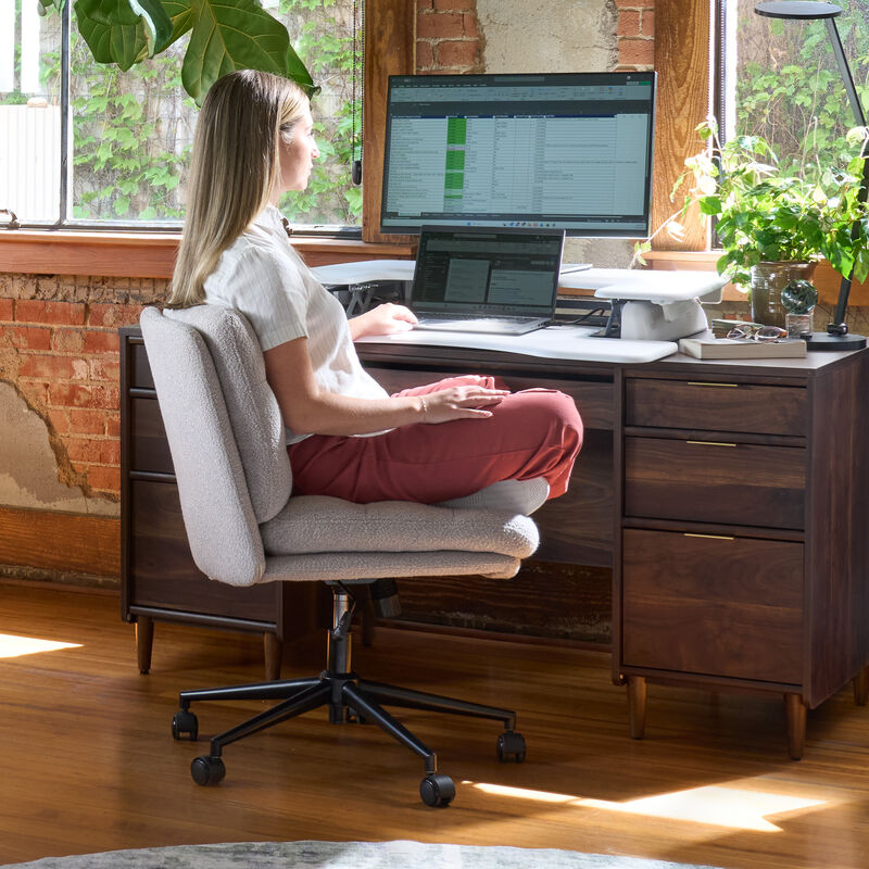 Woman sitting cross-legged in the light gray boucle armless office chair while working at a laptop on a raised desk converter, in a sunlit loft office with exposed brick. image number null