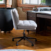 Side and back view of the light gray boucle armless office chair beside a large black planter and wood desk, showing the channel-tufted backrest and adjustable gas lift base.