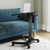 Mobile laptop desk with tilted black tabletop and laptop resting on top, shown beside a teal velvet sofa in a living room with a white shag rug, highlighting the tilt mechanism and height-adjustment crank.
