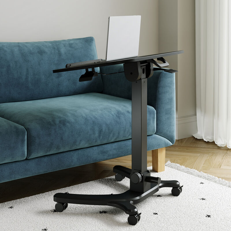 Mobile laptop desk with tilted black tabletop and laptop resting on top, shown beside a teal velvet sofa in a living room with a white shag rug, highlighting the tilt mechanism and height-adjustment crank. image number null