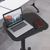 Overhead view of the desk's black tabletop holding an open laptop, a pen, and a brown leather notebook, showing the rounded edge design and built-in cable channel, with a white table and caster wheels visible in the background.