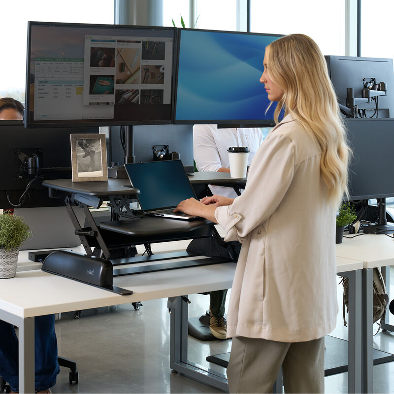 A professional office environment featuring a woman with long blonde hair using a black standing desk converter on a white desk. She is typing on a laptop, with two large monitors mounted above her workspace. The desk setup includes a framed photo, a small potted plant, and an ergonomic keyboard tray. In the background, other employees are working at similar standing desk setups, with modern office furnishings, large windows, and natural lighting creating a bright and collaborative atmosphere. image number null