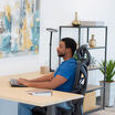 Side profile of a man in a blue shirt typing at a desk while seated in a black Vari ComfortFlex chair with headrest, in an office with shelving and plants.