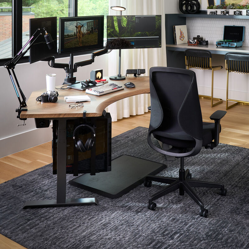 Modern home office setup featuring a black ergonomic Vari Tempur-Pedic chair at a curved wood desk with triple monitor arms, editing equipment, camera gear, and a gray area rug on hardwood flooring. image number null