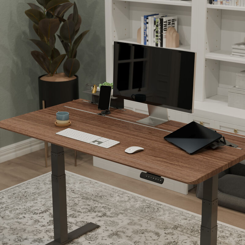 TechTrack ESD Walnut desk styled in a living room with a monitor, keyboard, mouse, tablet, phone stand, and mug, near a white bookshelf and a potted plant. image number null