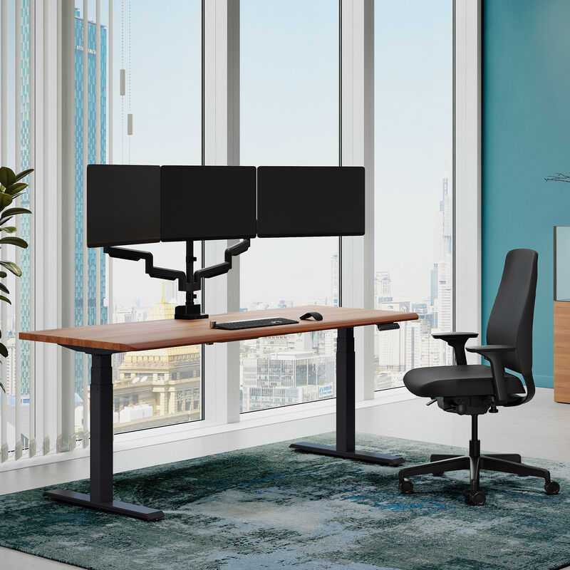 Solid Wood Electric Standing Desk with a walnut top and silver frame shown from a slightly different angle in the same sunlit home office, with a triple-monitor arm mounted above the desktop. image number null