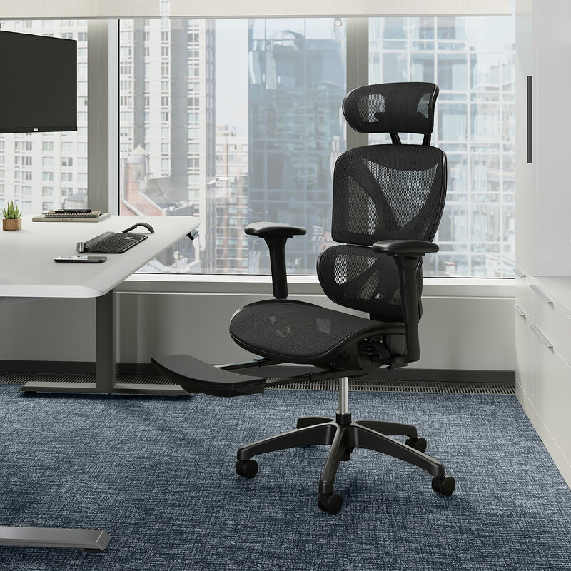 Black mesh Vari ComfortFlex chair with headrest, adjustable arms, and extended footrest in a modern office with floor-to-ceiling windows and city view, beside a white desk. image number null