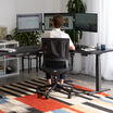 Man seated in a black Vari Essential Task Chair working at a triple-monitor desk setup, viewed from behind in a home office with a striped area rug.