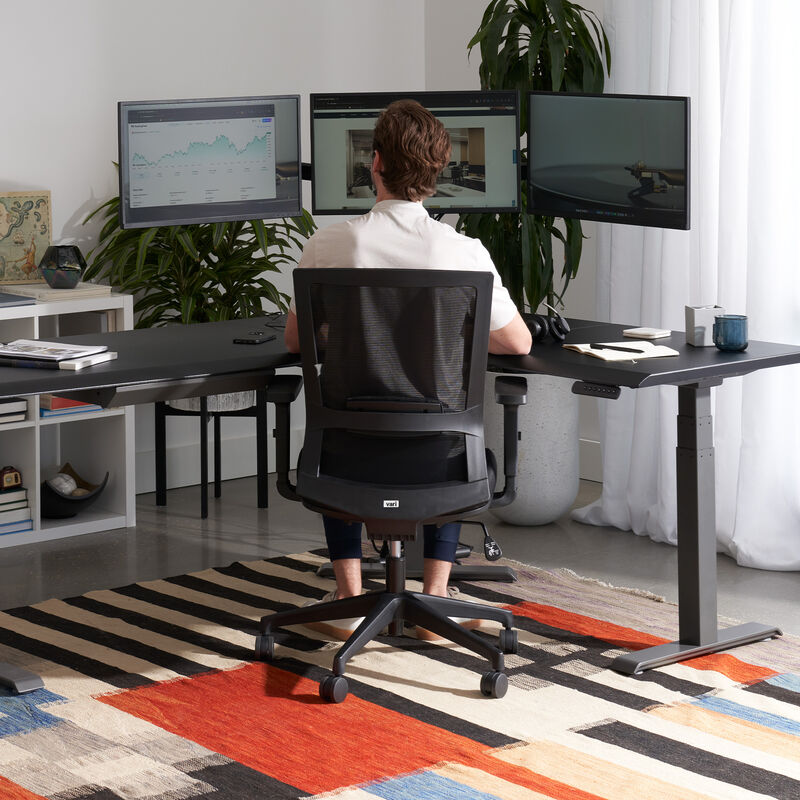 Man seated in a black Vari Essential Task Chair working at a triple-monitor desk setup, viewed from behind in a home office with a striped area rug. image number null