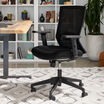 Front angled view of black Vari Essential Task Chair with mesh backrest, cushioned seat, and adjustable armrests, in an office with bookshelves and a plant.