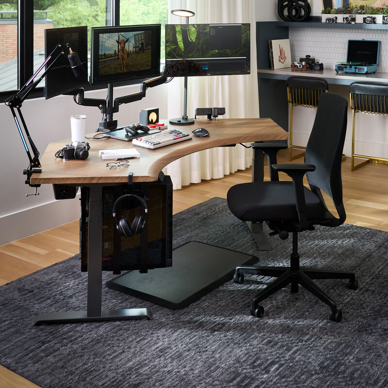 Modern home office setup featuring a black ergonomic Vari Tempur-Pedic chair at a curved wood desk with triple monitor arms, editing equipment, camera gear, and a gray area rug on hardwood flooring. image number null