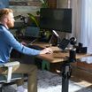 Man working at a walnut TechTrack ESD standing desk fitted with TechTrack accessories including a monitor arm, tablet mount, phone mount, speakers, cup holder, headphone hook, and cable management rail, in a home office with exposed brick and plants.