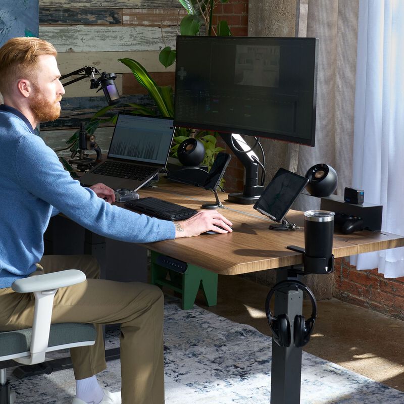 Man working at a walnut TechTrack ESD standing desk fitted with TechTrack accessories including a monitor arm, tablet mount, phone mount, speakers, cup holder, headphone hook, and cable management rail, in a home office with exposed brick and plants. image number null