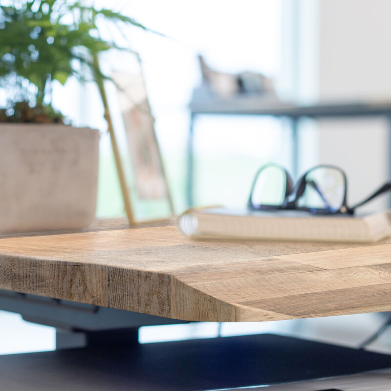 Close-up of the beveled edge of a reclaimed wood-look desktop, with a notebook, glasses, picture frame, and potted plant blurred in the background. image number null