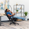 Man in a blue shirt reclining in a black Vari ComfortFlex chair with headrest and extended footrest, talking on the phone in an office with a desk and shelving.