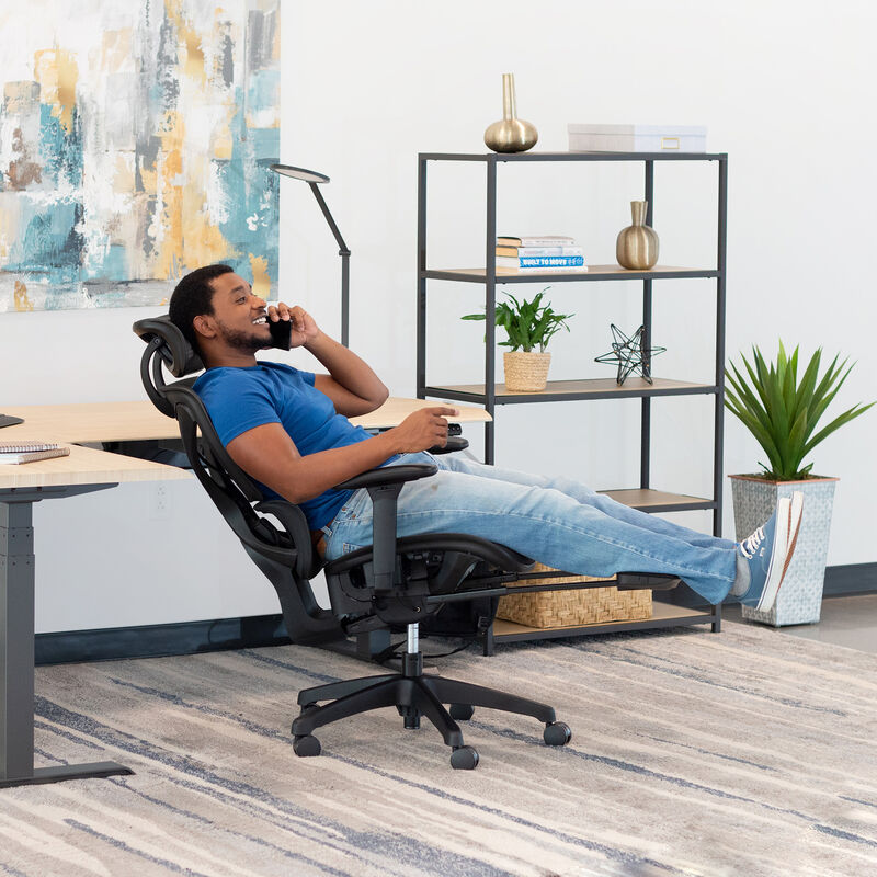 Man in a blue shirt reclining in a black Vari ComfortFlex chair with headrest and extended footrest, talking on the phone in an office with a desk and shelving. image number null