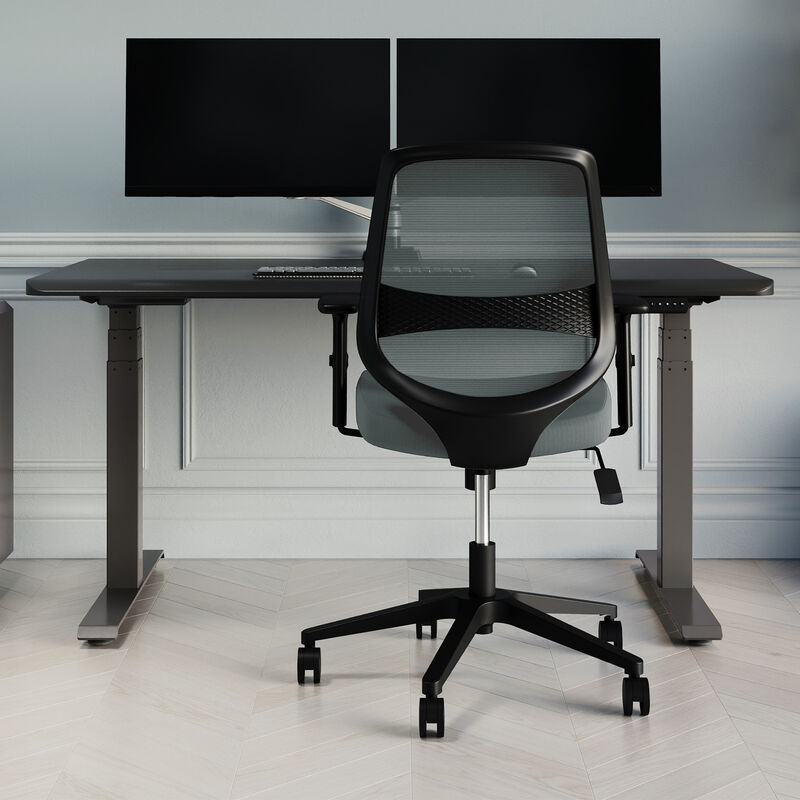 Rear three-quarter view of the sage grey mesh office chair showing the black back panel and five-star base with casters, positioned beside a sit-stand desk with dual monitors. image number null