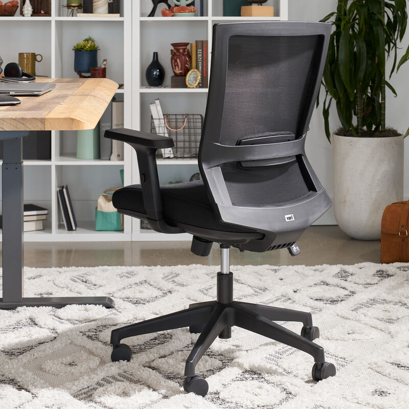 Rear view of black Vari Essential Task Chair with mesh backrest and adjustable armrests, positioned beside a wood desk in an office with bookshelves and a plant image number null