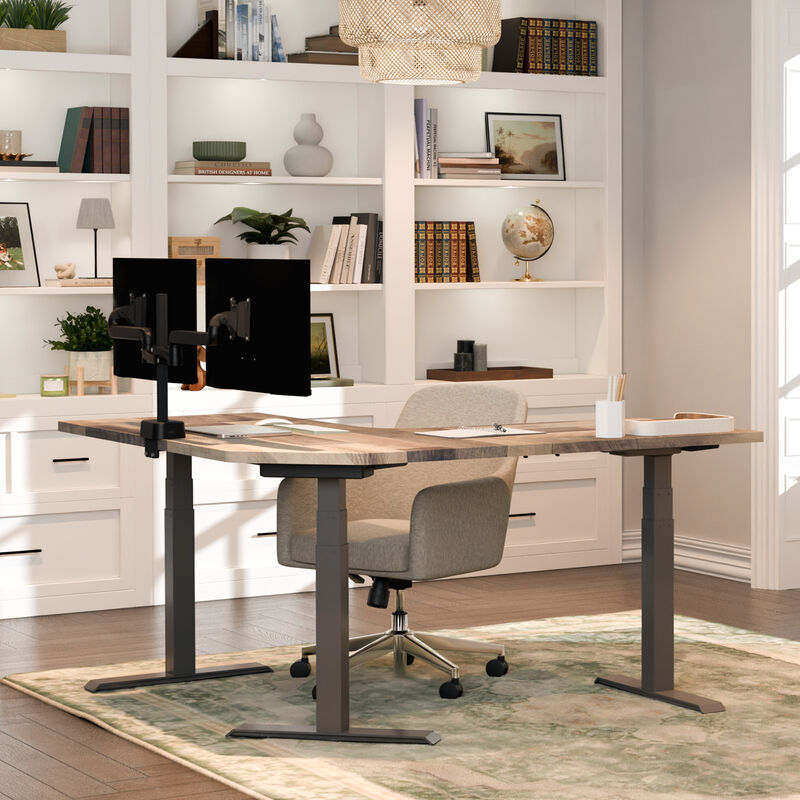 L-shaped standing desk with a reclaimed wood-look top and dark gray frame, holding a dual monitor arm, laptop, and notebook, in a home office with white built-in shelves and a beige chair. image number null