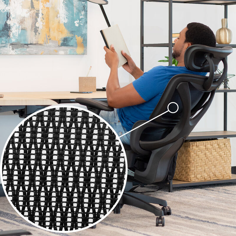 Man in a blue shirt reading a book while reclining in a black Vari ComfortFlex chair, with a close-up circular inset showing the woven mesh backrest texture. image number null