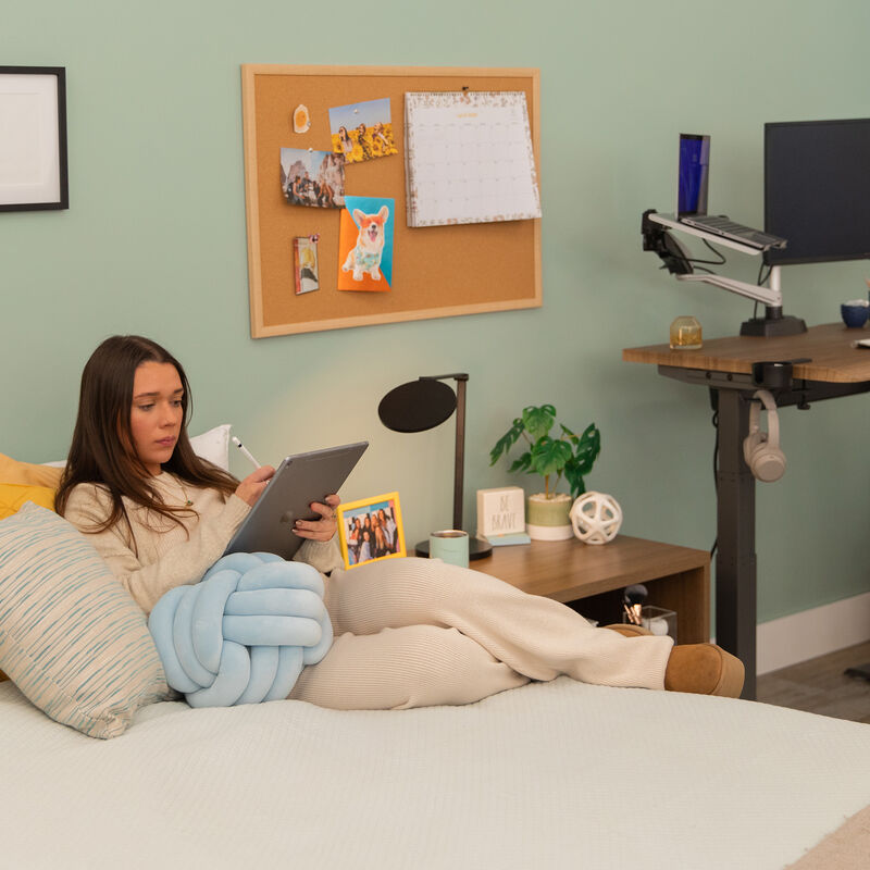 A woman lounges on a bed using a tablet in a cozy home office with a Vari Studio Lamp on the bedside table. image number null