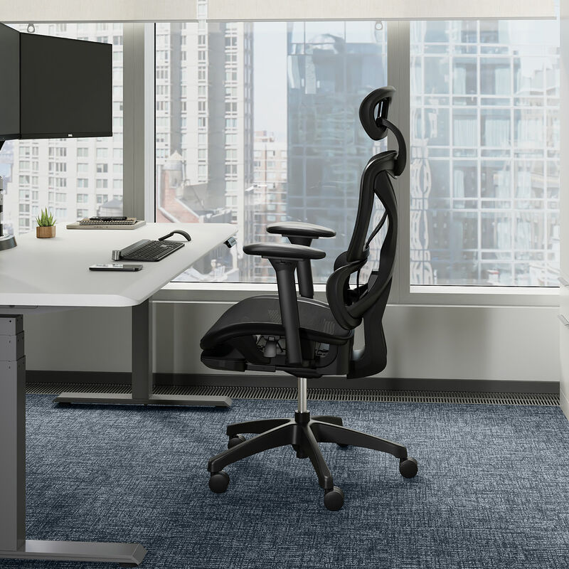 Side view of black mesh Vari ComfortFlex chair with headrest, adjustable armrests, and retracted footrest, positioned beside a white desk near floor-to-ceiling windows. image number null