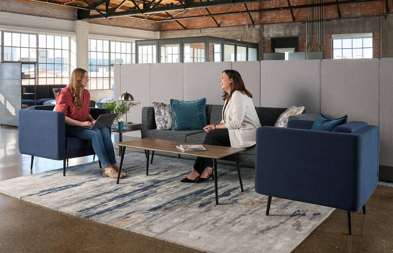 two women sitting on a sofa and side chair working on their laptops in an open office setting