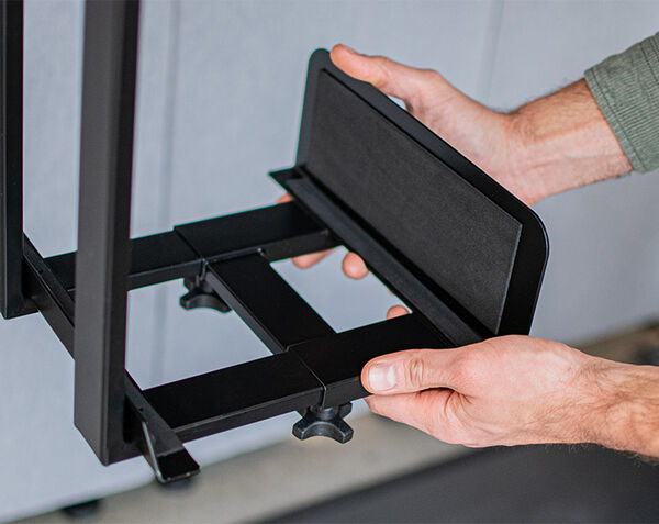 Person adjusting a black metal desk mount platform with padded sides, to hold the PC case, using both hands.