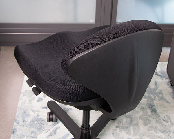 Close-up of black office chair seat and backrest from behind, showing contoured split seat design, black frame, and caster wheels on an area rug.