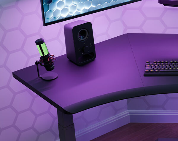 Corner of the black Command Center Electric Standing Desk under purple lighting, with a green-lit microphone on a stand, a speaker, and a mechanical keyboard on the surface, plus a monitor mounted above.