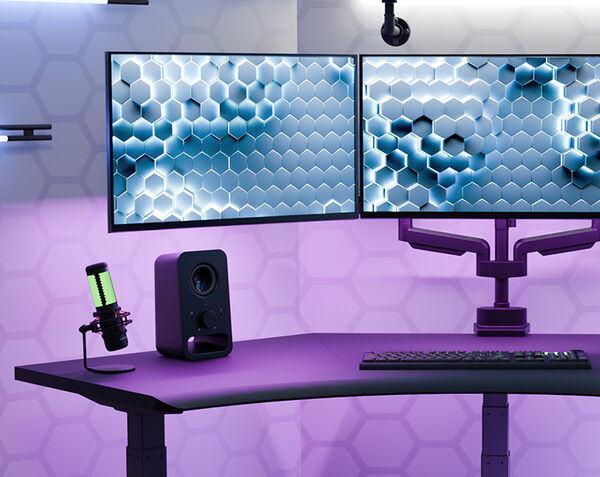 Wide view of the black Command Center Electric Standing Desk under purple lighting, set up with dual monitors on an arm, a green-lit microphone, a speaker, and a mechanical keyboard on the surface.