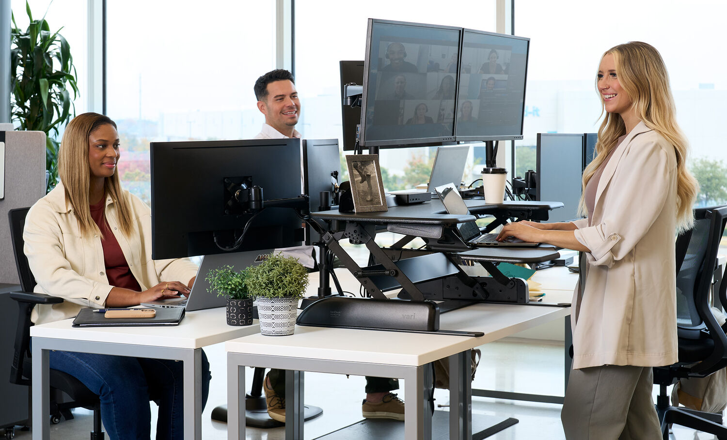 A collaborative office environment with three professionals working at modern desks equipped with dual-monitor setups and standing desk converters. A woman with long blonde hair is standing and typing on her laptop, while another woman with shoulder-length hair is seated at an adjacent desk, working on her computer. A man in the background, wearing a white shirt, is smiling and engaged in conversation. The desks are decorated with potted plants, framed photos, and office essentials, while large windows allow natural light to brighten the space.