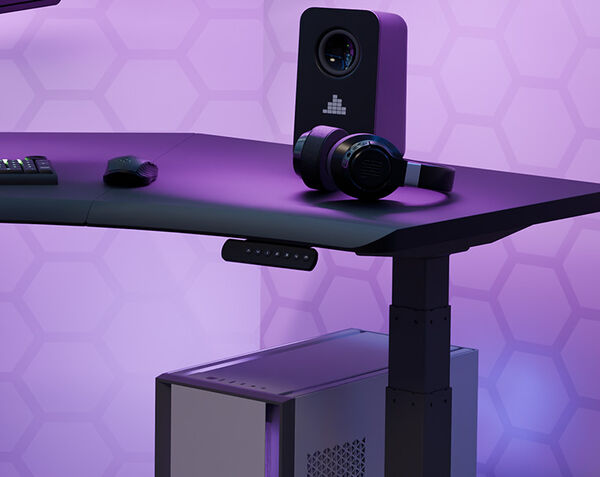 Corner of the black Command Center Electric Standing Desk under purple lighting, with a speaker and headphones resting on the surface, a keyboard and mouse nearby, a control panel on the front edge, and a computer tower underneath.