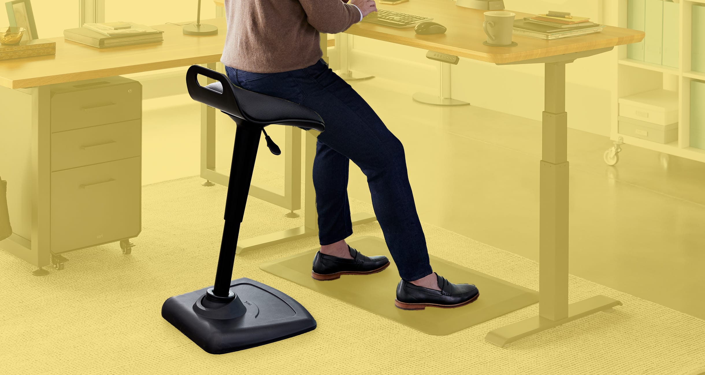 individual sitting on a stool leaning and working at an electric standing desk