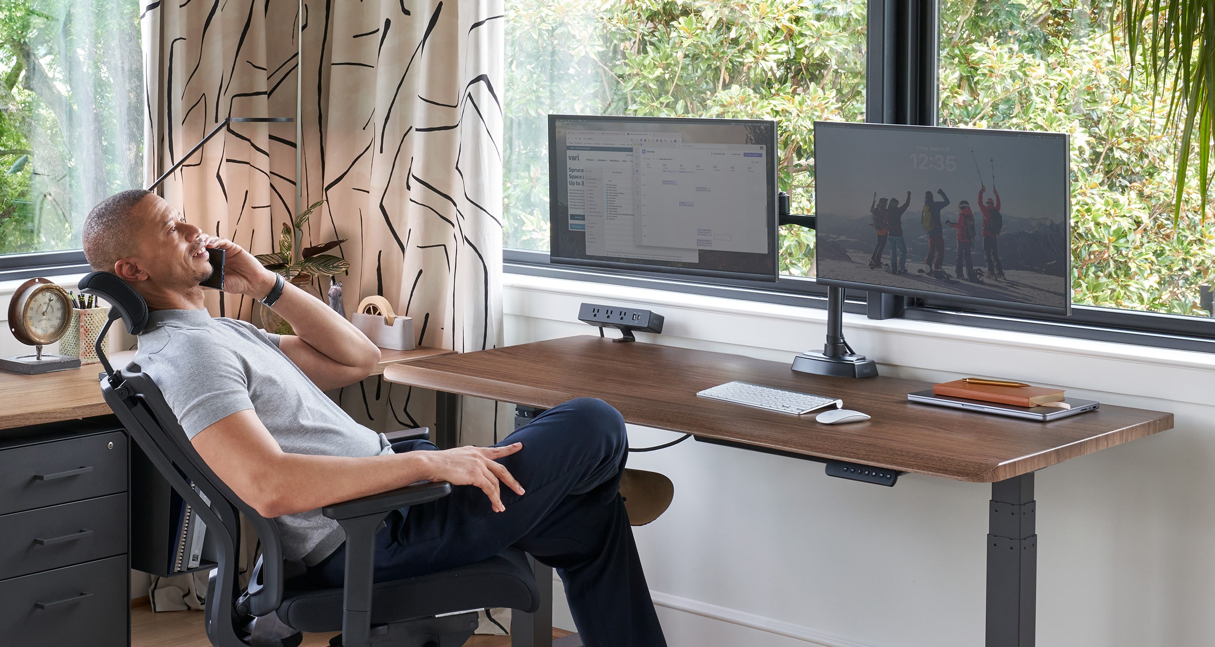 a man sitting in an office chair leaning back talking on the phone in front of an electric standing desk in a home office setting