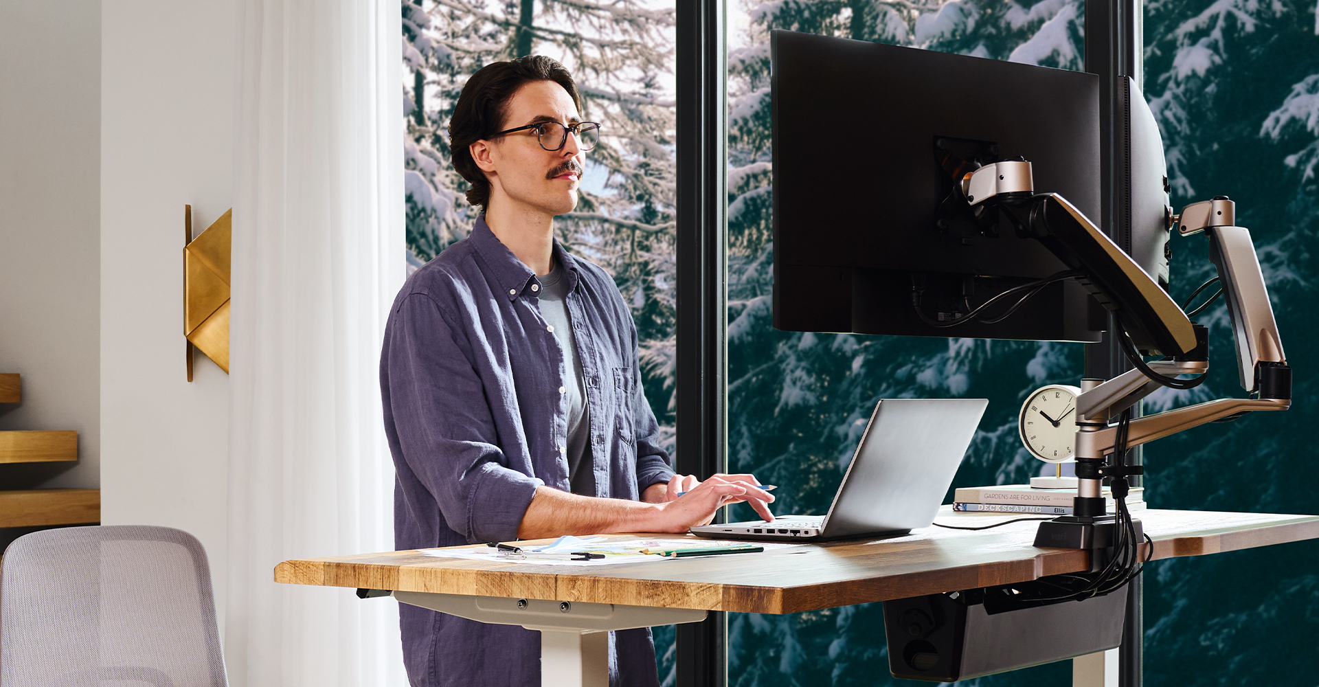man standing working at an electric standing desk in front of a window in a home office area